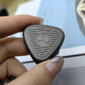 A hand with manicured nails holds a gray EDC Fingertip Toys Fidget Slider—triangular, metallic, with engraved lines and a central circular icon. The slightly blurred background emphasizes the toys sleek finish.