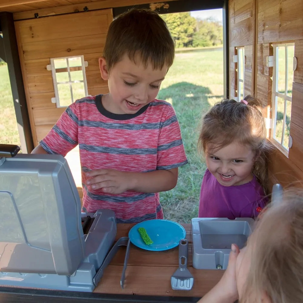 Two young children play together in their Playhouse Backyard Discovery Canyon Creek Cedar Swing Set, pretending to use a toy cash register and kitchen set, smiling and having fun outdoors on a sunny day.
