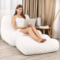 A woman in a white dress sits barefoot on the Widen-Cream Bean Bag Sofa with Footstool, using her laptop. Her legs are stretched out in a modern room with wooden floors, curtains, and a small bookshelf.