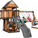 Children play on the Playhouse Backyard Discovery Canyon Creek Cedar Swing Set with a grey slide, swings, climbing wall, clubhouse, and picnic table. Some kids swing, slide, and climb together outdoors enjoying this fun playset.