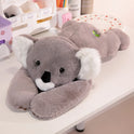 A Long Arm Animal Weighted Plush Soft Pillow in Koala color lies on a white desk, surrounded by office supplies and decor. The plush features soft gray fur with white ears and a black nose.