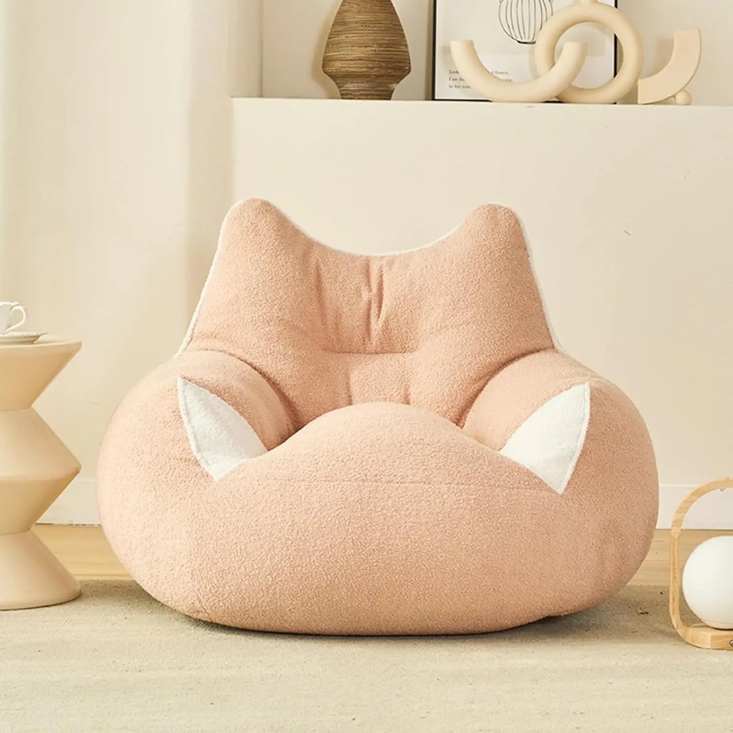 A Premium Teddy Velvet Bean Bag Sofa Cover in pink, featuring white ear-like details and extra storage for plush toys, adds a soft touch to a cozy room with minimalist decor and a small side table.