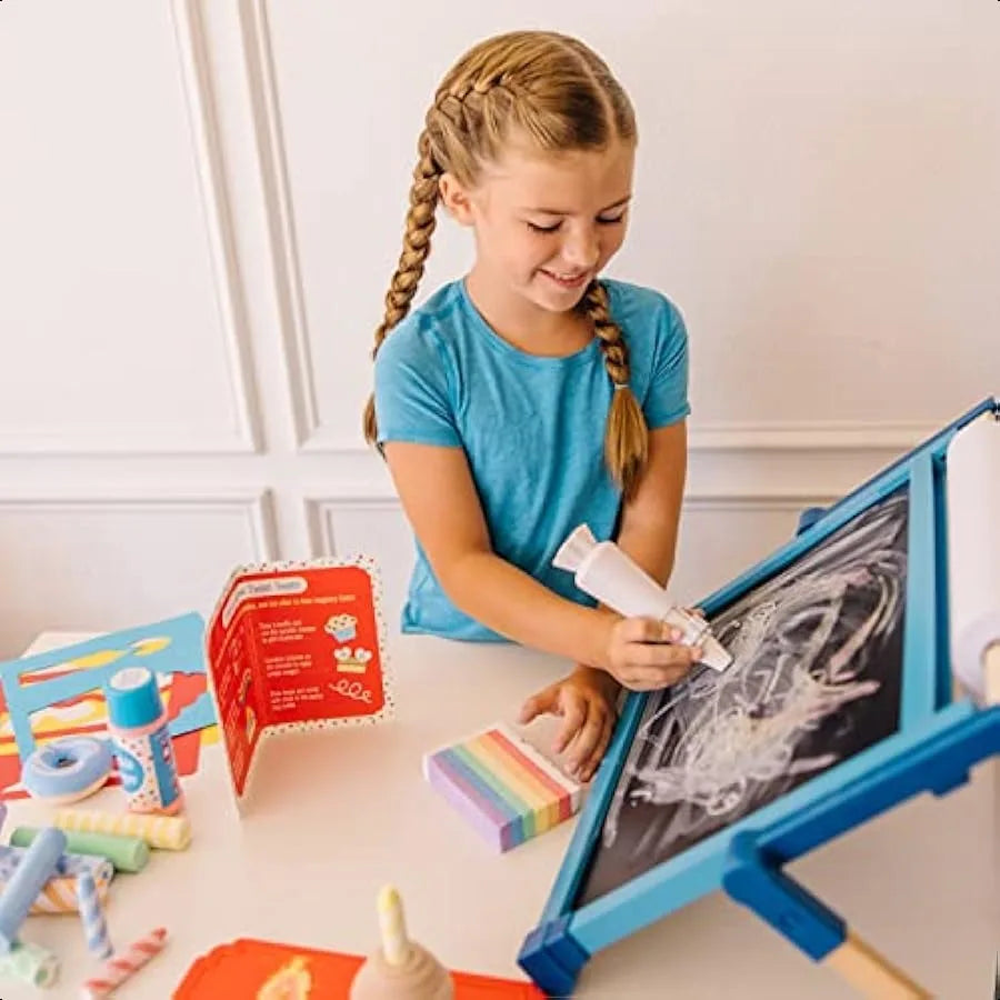 A young girl with braided hair smiles as she draws on a black chalkboard easel using the Melissa and Doug Sweet Shop Multicolored Chalk and Holders Play Set (33 Pieces); safe art supplies and colorful toys are on the table beside her.