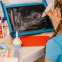 A child with braided hair draws an animal with white chalk from the Melissa and Doug Sweet Shop Multicolored Chalk and Holders Play Set on a small blackboard, while toy cupcakes and art supplies are scattered on the white table nearby.