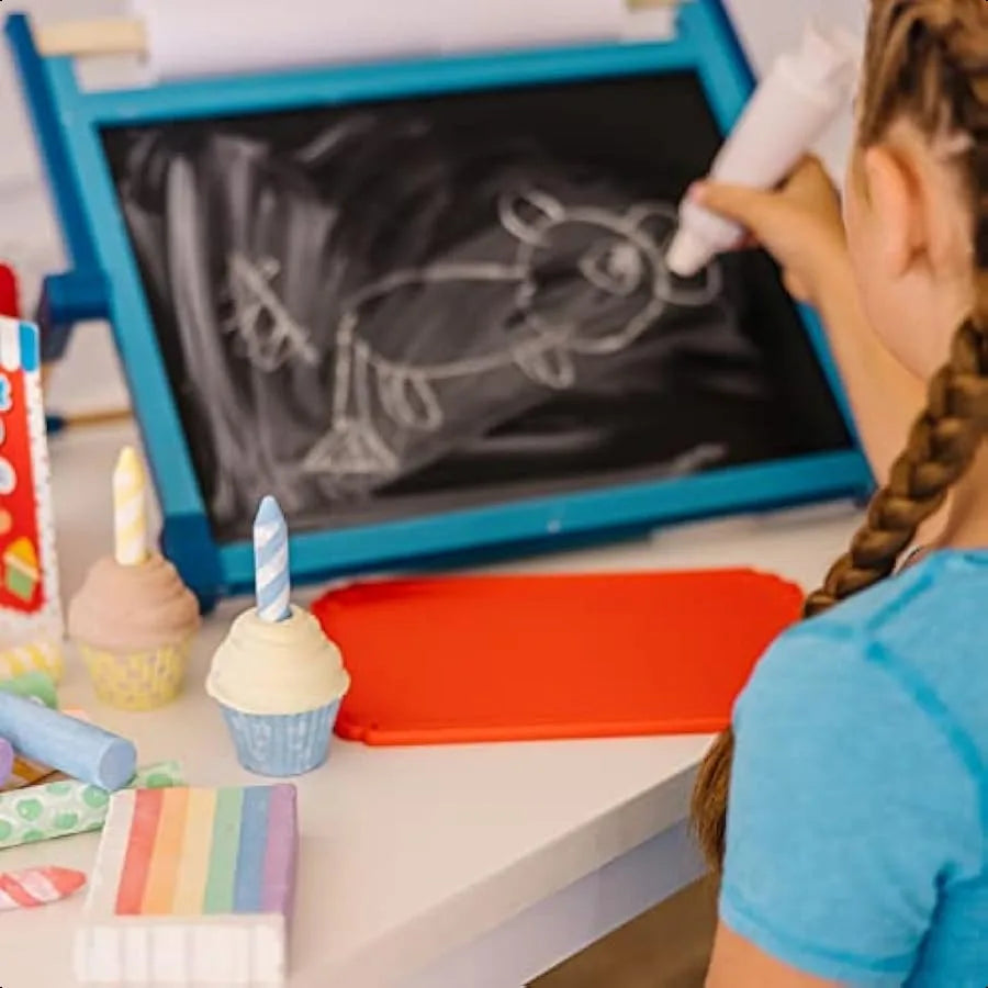 A child with braided hair draws an animal with white chalk from the Melissa and Doug Sweet Shop Multicolored Chalk and Holders Play Set on a small blackboard, while toy cupcakes and art supplies are scattered on the white table nearby.