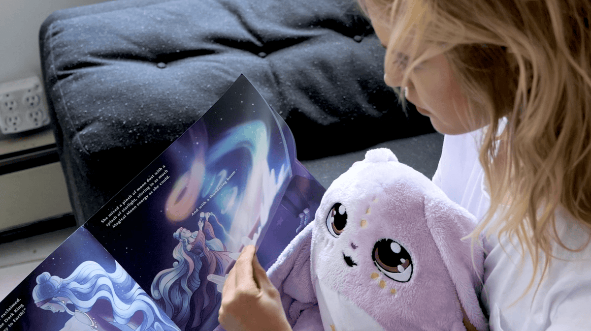 A person reading a Moon Pals Picture Book to a plush lavender creature with large, expressive eyes.