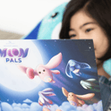 A child reads a Moon Pals Picture Book featuring colorful, whimsical Moon Pals flying against a backdrop of the moon and stars.