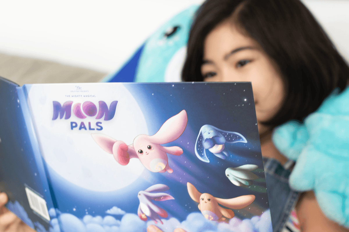 A child reads a Moon Pals Picture Book featuring colorful, whimsical Moon Pals flying against a backdrop of the moon and stars.