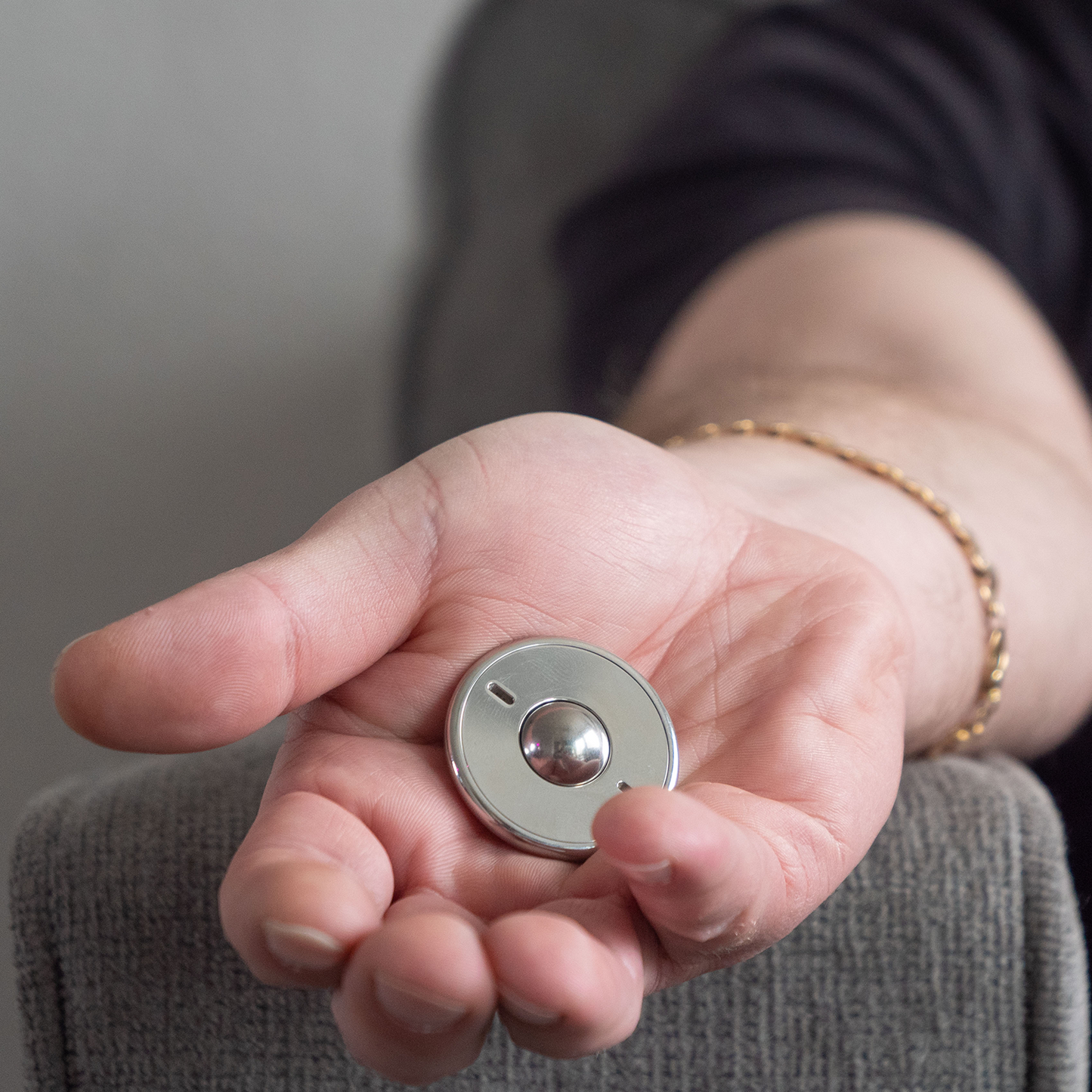 A person with a gold bracelet holds an ONO Scroller Steel fidget toy in their open hand while resting their arm on a grey upholstered chair.