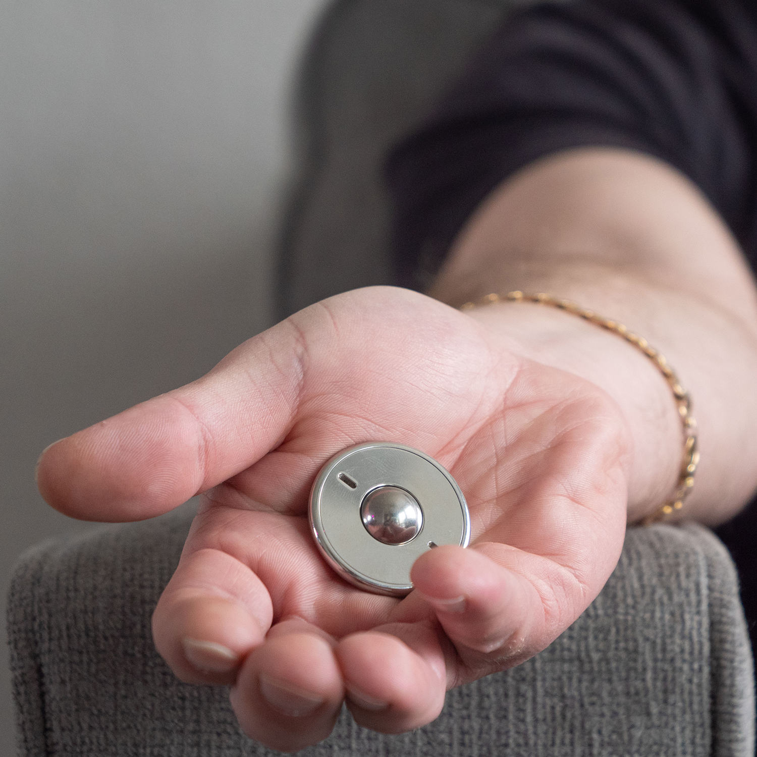 A person with a gold bracelet holds an ONO Scroller Steel fidget toy in their open hand while resting their arm on a grey upholstered chair.
