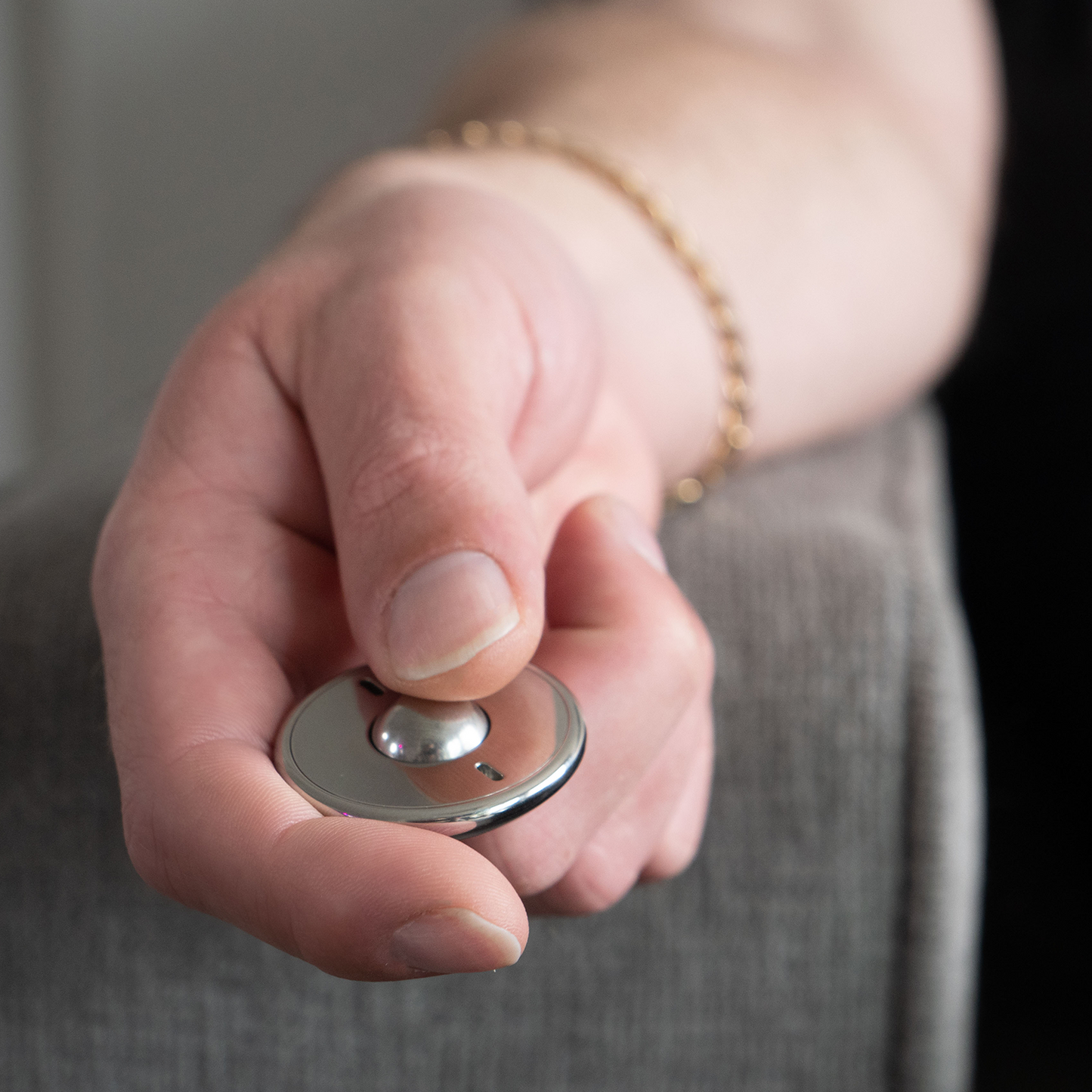 A hand wearing a gold bracelet holds the small, round ONO Scroller Steel remote on the arm of a gray upholstered chair.