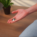 A person holds a sleek ONO Scroller Steel in their palm, with a green potted plant and wooden table in the background.