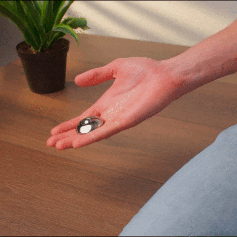 A person holds a sleek ONO Scroller Steel in their palm, with a green potted plant and wooden table in the background.
