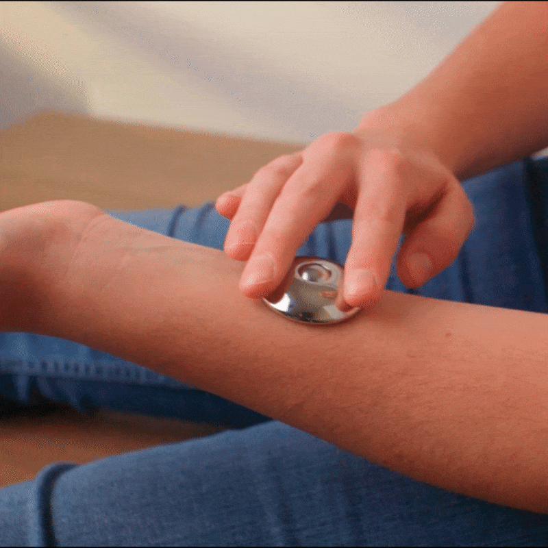 Someone rolls an ONO Scroller Steel over their forearm, likely for soothing or cooling. In the background, part of their blue jeans and a beige surface are visible.