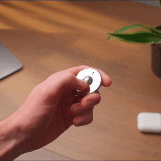 A hand spins an ONO Scroller Steel metallic fidget toy over a wooden desk with a laptop, plant, and wireless earbuds in the background.
