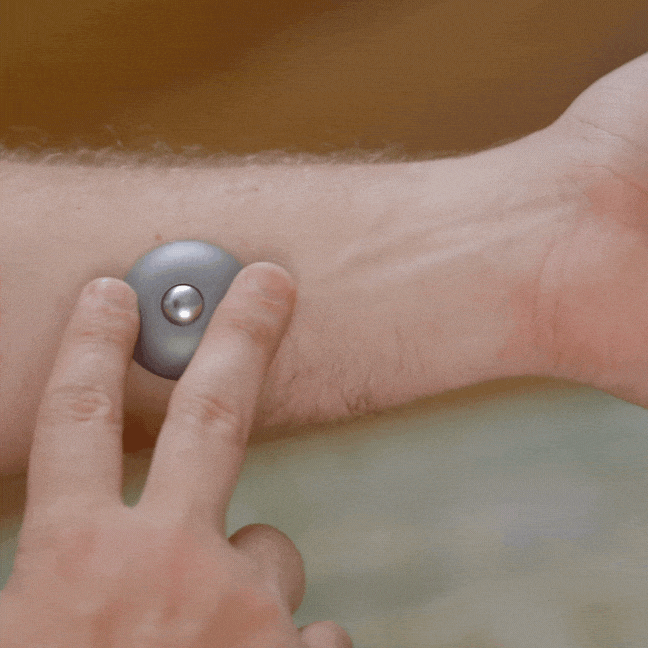 A person presses an ONO Scroller Titanium—small, round, and gray—against the inside of their arm, showing how it works on bare skin.
