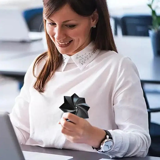 A woman with long brown hair, wearing a white blouse and a black geometric Fidget 3D Spinner Stress Relief Toy brooch, sits at her desk smiling and working on a laptop in an office setting.