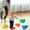 A toddler in a white shirt and dark pants navigates the Kids Balance River Stones - 5 Non-Slip Sensory Toys. An adult crouches nearby, smiling supportively as sunlight streams through windows, highlighting this playful obstacle course.