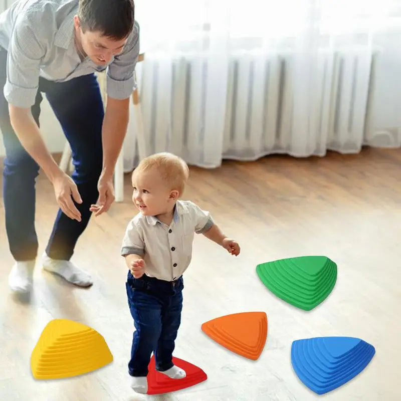 A toddler in a white shirt and dark pants navigates the Kids Balance River Stones - 5 Non-Slip Sensory Toys. An adult crouches nearby, smiling supportively as sunlight streams through windows, highlighting this playful obstacle course.