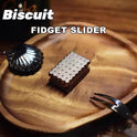 The Biscuits Fidget Slider, a metal haptic toy with a biscuit cartoon design and rustproof build, sits on a wooden tray beside a black shell-shaped dish and fork. Biscuit Fidget Slider is displayed at the top of the image.