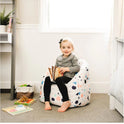 A young blonde-haired child with a black bow sits on the Milano Kids Bean Bag Chair for Comfort and Fun, holding an open book. Toys and books are scattered nearby as sunlight streams through the window behind her.