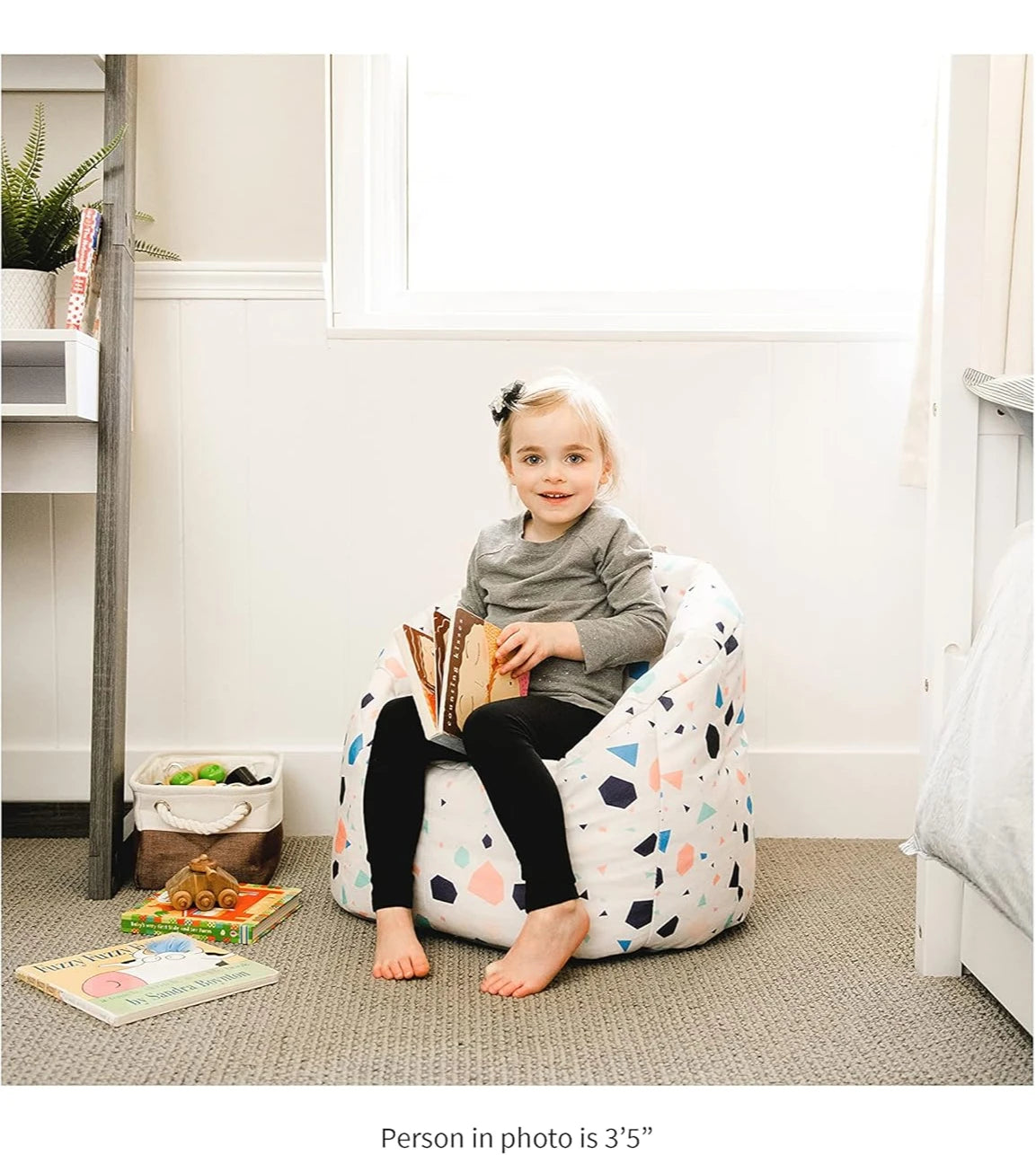 A young blonde-haired child with a black bow sits on the Milano Kids Bean Bag Chair for Comfort and Fun, holding an open book. Toys and books are scattered nearby as sunlight streams through the window behind her.