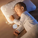 A baby in white pajamas sleeps peacefully on a bed, comforted by the Portable Sleep Aid Device for Baby with 10 soothing sounds and a creative night light, perfect for restful sleep in the crib or while traveling.