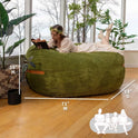 A woman in a light dress and flower crown reads on a Fuf XXL Foam Filled Bean Bag Chair, Moss Green, 6 Feet, lounging in a sunlit room with plants and large windows. (Dimensions: 71W x 58D x 39H.).