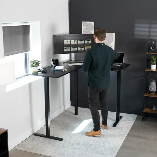 A man works at an Electric Height Adjustable 63 x 55 Inch Corner Stand Up Desk in a modern office with dual monitors, a laptop, lamp, and decor. Natural light shines through a window into the workspace.