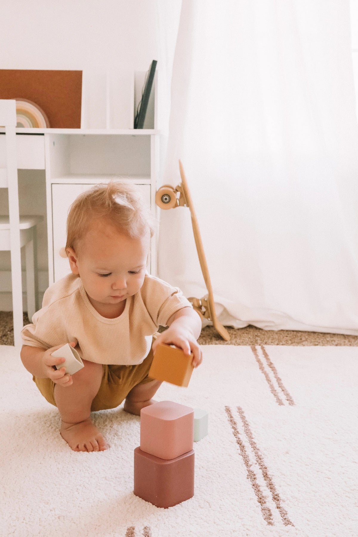 A toddler sits on a rug stacking five colorful, square-shaped Silicone Stacking Cups (Set of 5).