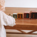 A child reaches toward five Silicone Stacking Cups (Set of 5) arranged in a row on a wooden table, each containing a small peg person.