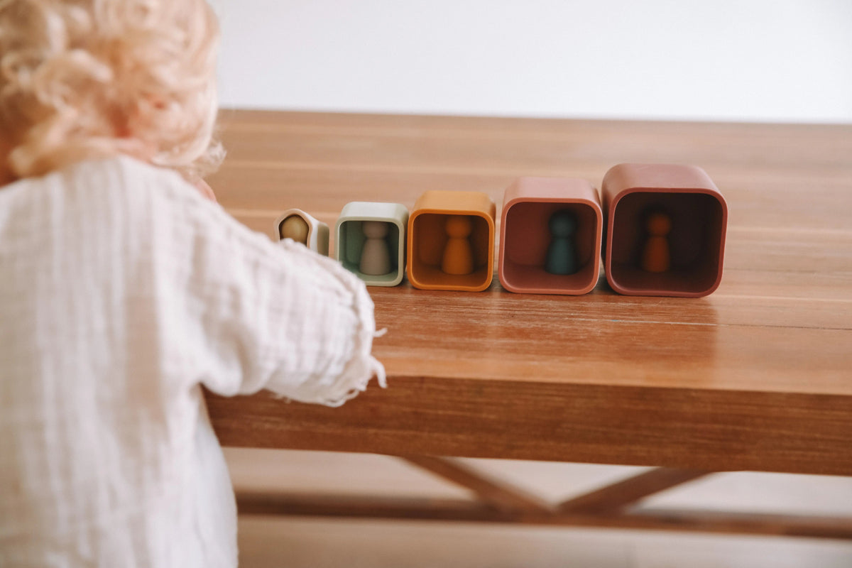 A child reaches toward five Silicone Stacking Cups (Set of 5) arranged in a row on a wooden table, each containing a small peg person.