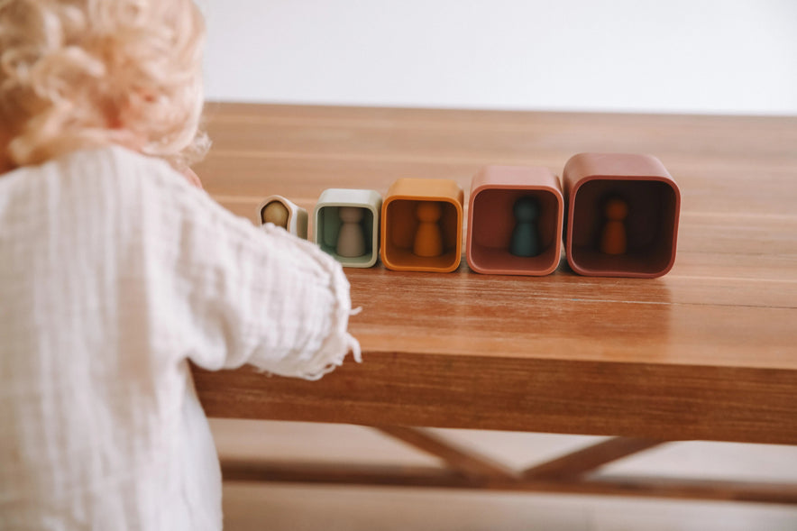 A child reaches toward five Silicone Stacking Cups (Set of 5) arranged in a row on a wooden table, each containing a small peg person.