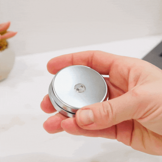 A hand holds an ONO Slider Steel, flicking the sleek steel fidget slider—this modern stress-relief tool stands out against a blurred plant and part of a laptop in the background.