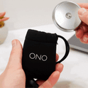 A hand pulls a round silver ONO Slider Steel fidget toy from a small black drawstring pouch, with a plant and a laptop in the background.