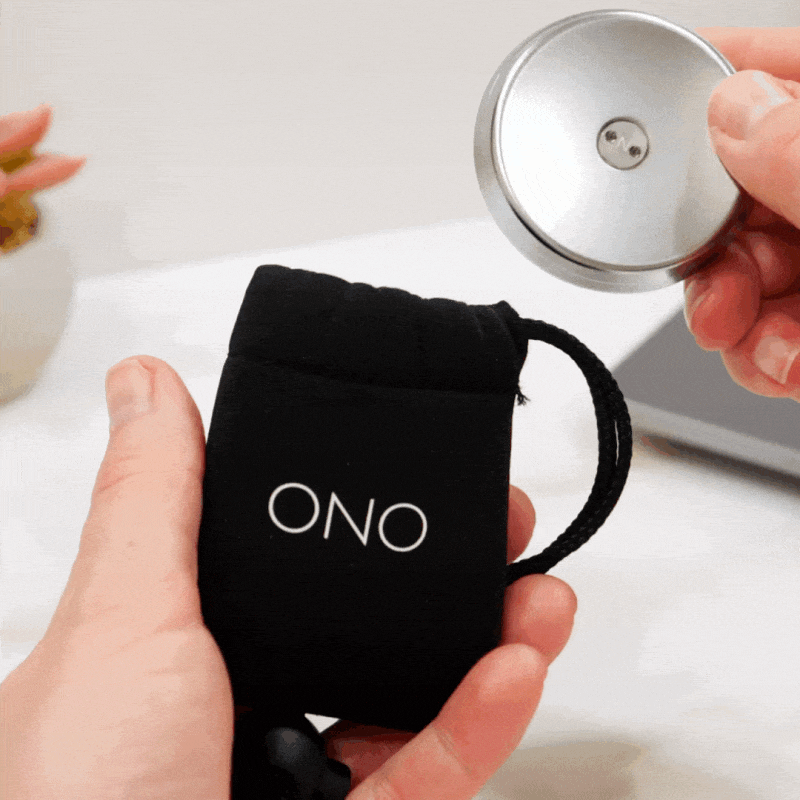 A hand pulls a round silver ONO Slider Steel fidget toy from a small black drawstring pouch, with a plant and a laptop in the background.