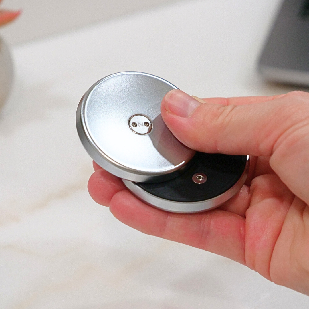 A hand holds and twists the ONO Slider Steel, a round, silver and black metallic fidget toy with visible small screws, on a white surface.