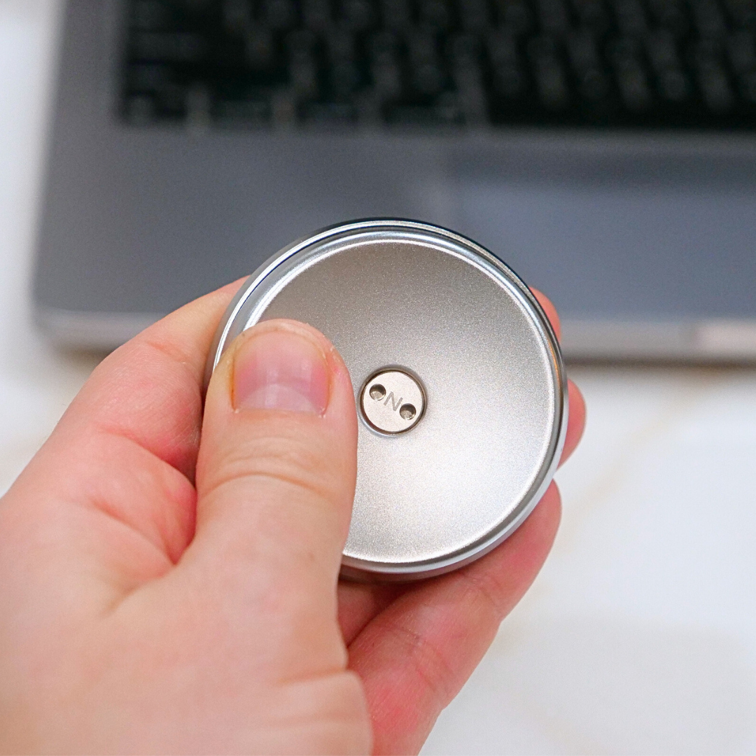 A hand holds a small, round ONO Slider Steel fidget toy with ONO engraved in the center, in front of a blurred laptop keyboard.