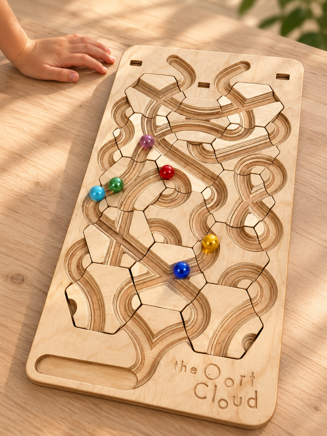 A child’s hand reaches toward The Oort Cloud – Wooden Marble Run Puzzle, which features carved tracks and several colorful glass marbles.