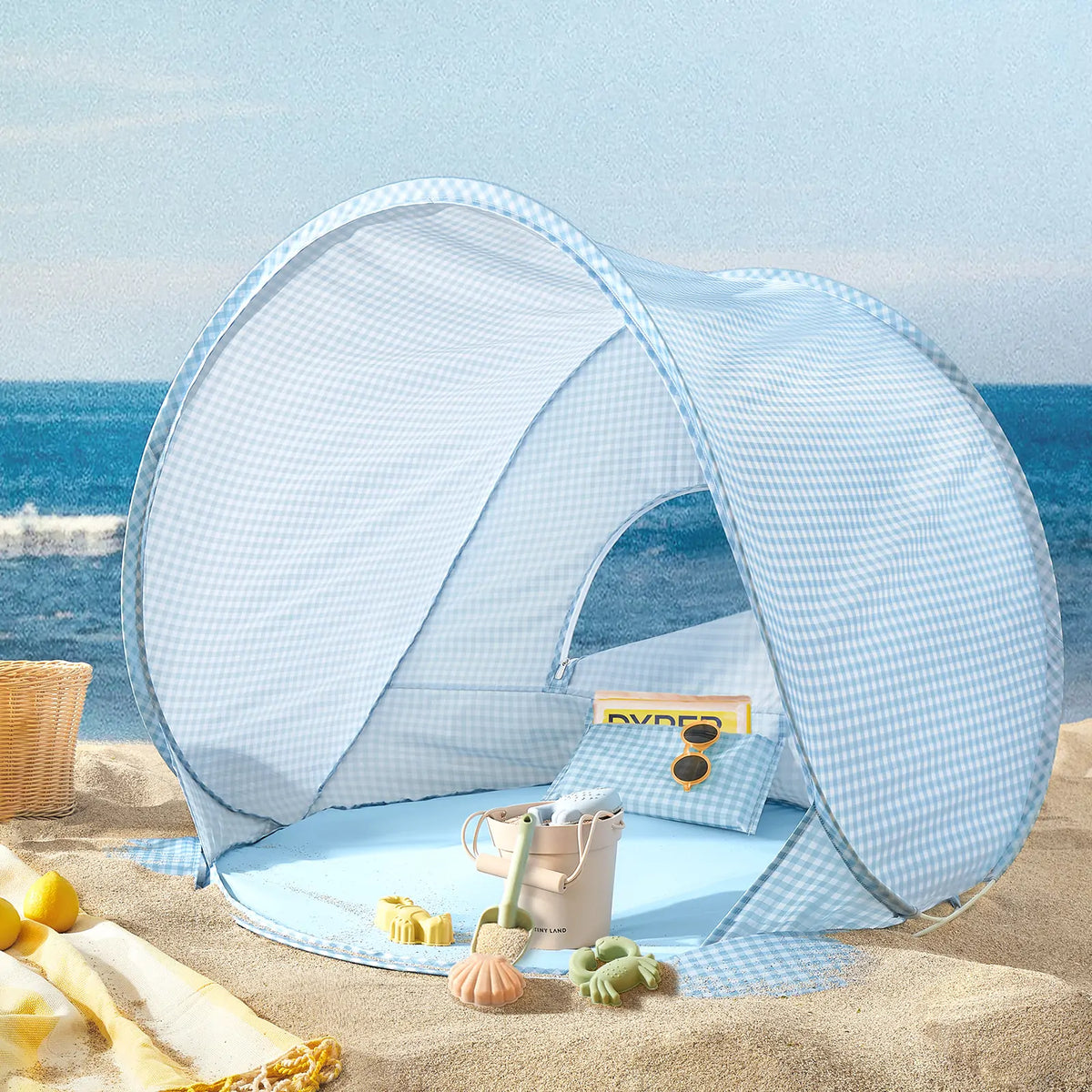 Light blue gingham Kids Beach Tent & Sand Toy Set on the sand, containing a bucket, a shovel, and a sand toy set.