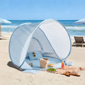 A blue-and-white gingham Kids Beach Tent & Sand Toy Set on the sand, containing a small pillow, sunglasses, and molds, with a plate of grapes and sandals nearby.