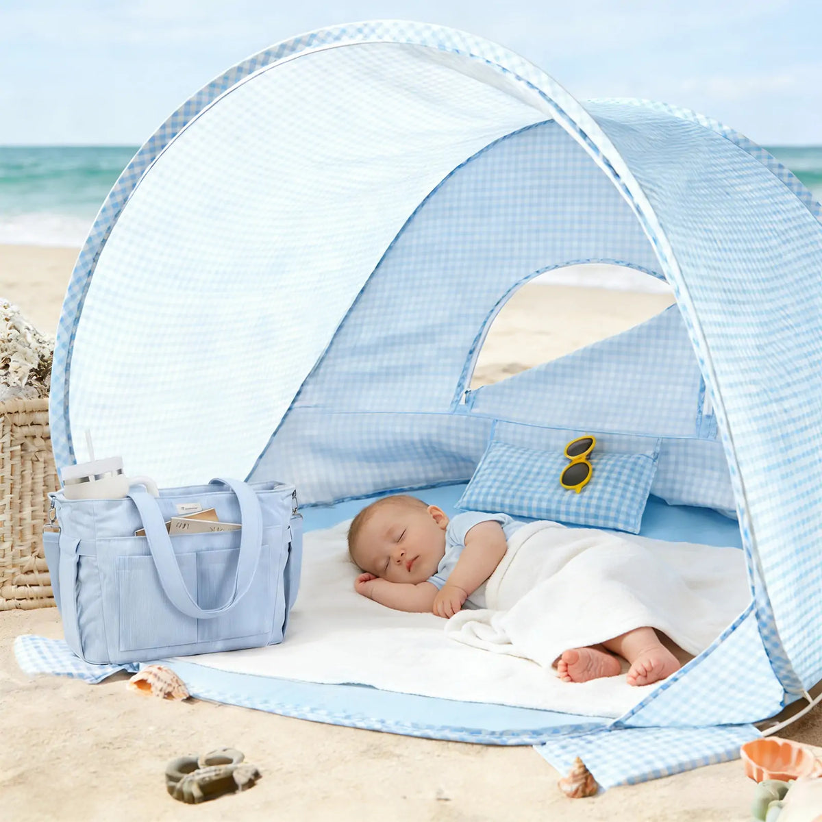 A baby sleeps under a blue and white checkered Kids Beach Tent & Sand Toy Set, with a matching tote bag, sunglasses, and the toy set placed nearby on the sand.