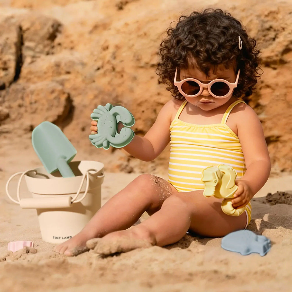 A toddler in sunglasses and a striped swimsuit plays with a Kids Beach Tent & Sand Toy Set, using its bucket, shovel, and animal-shaped molds in the sand.