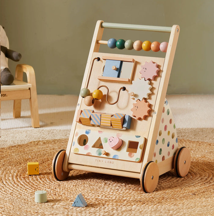 The Tiny Land® Versatile Natural Wooden Baby Walker, featuring colorful beads, gears, blocks, interactive shapes, and polka dot patterns on the sides, sits on a woven rug in a cozy, softly lit room.