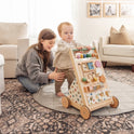 A toddler stands holding the Tiny Land® Versatile Natural Wooden Baby Walker as a smiling woman kneels behind, supporting the child in a cozy living room with a sofa, pillows, and framed photos on the wall.