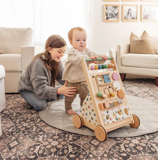 A toddler stands holding the Tiny Land® Versatile Natural Wooden Baby Walker as a smiling woman kneels behind, supporting the child in a cozy living room with a sofa, pillows, and framed photos on the wall.