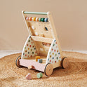 The Tiny Land® Versatile Natural Wooden Baby Walker with colorful beads, a shape sorter, and wheels stands on a woven rug. Pastel blocks and shapes—ideal for sensory development—are scattered nearby.