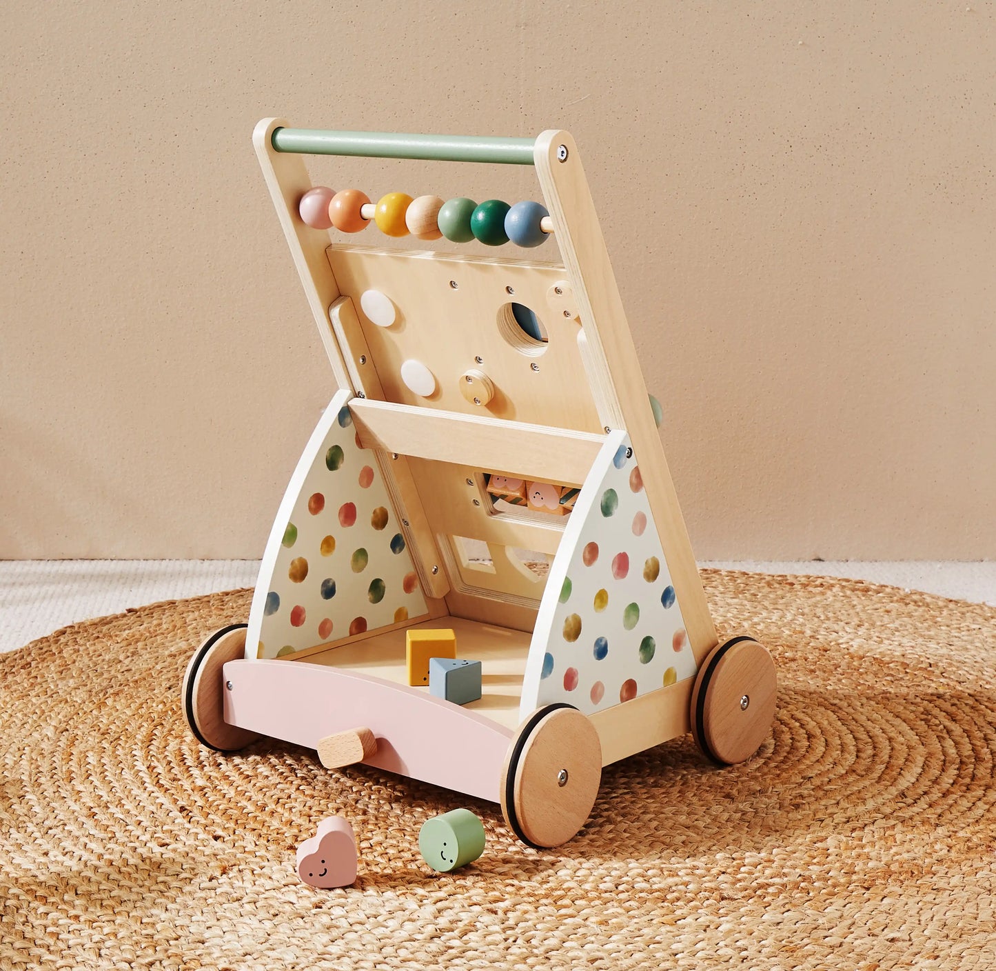 The Tiny Land® Versatile Natural Wooden Baby Walker with colorful beads, a shape sorter, and wheels stands on a woven rug. Pastel blocks and shapes—ideal for sensory development—are scattered nearby.