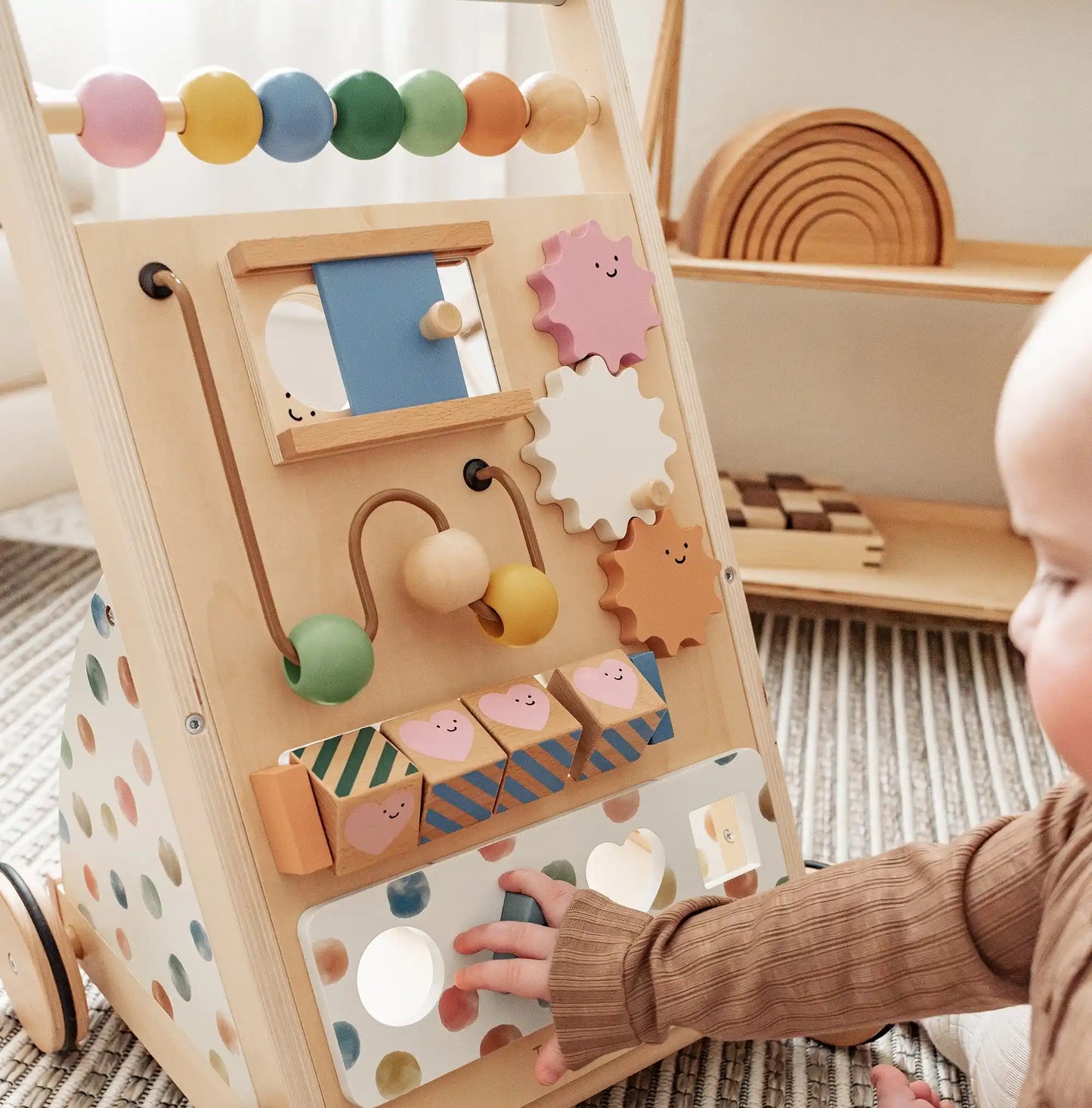 A baby plays with the Tiny Land® Versatile Natural Wooden Baby Walker—featuring colorful beads, gears, mirrors, and smiling blocks—an engaging sensory development toy on a striped rug with shelves and wooden toys behind.