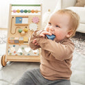 A smiling toddler in a brown shirt and gray pants sits on a mat, chewing a blue block, with the Tiny Land® Versatile Natural Wooden Baby Walker—featuring colorful beads and gears—placed behind them for sensory play.
